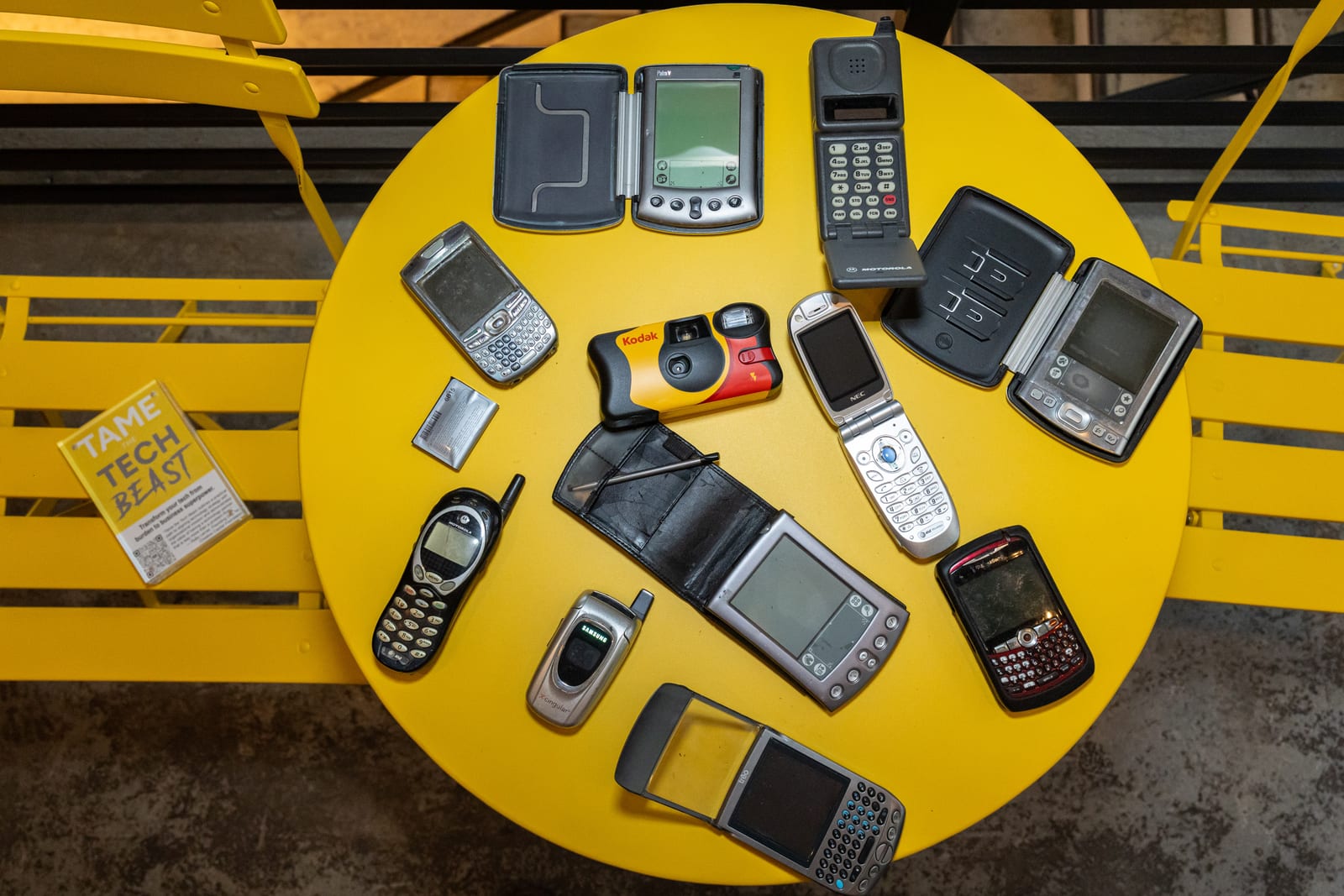 An overhead view of a yellow table covered in 2000s-era devices: PDAs, flip phones, a BlackBerry, and a yellow Kodak disposable camera