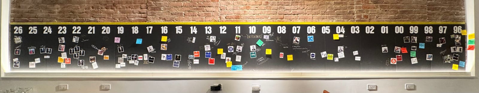 A wide view of a brick wall timeline running from 1996 to 2026, covered in photos, sticky notes, and handwritten milestones