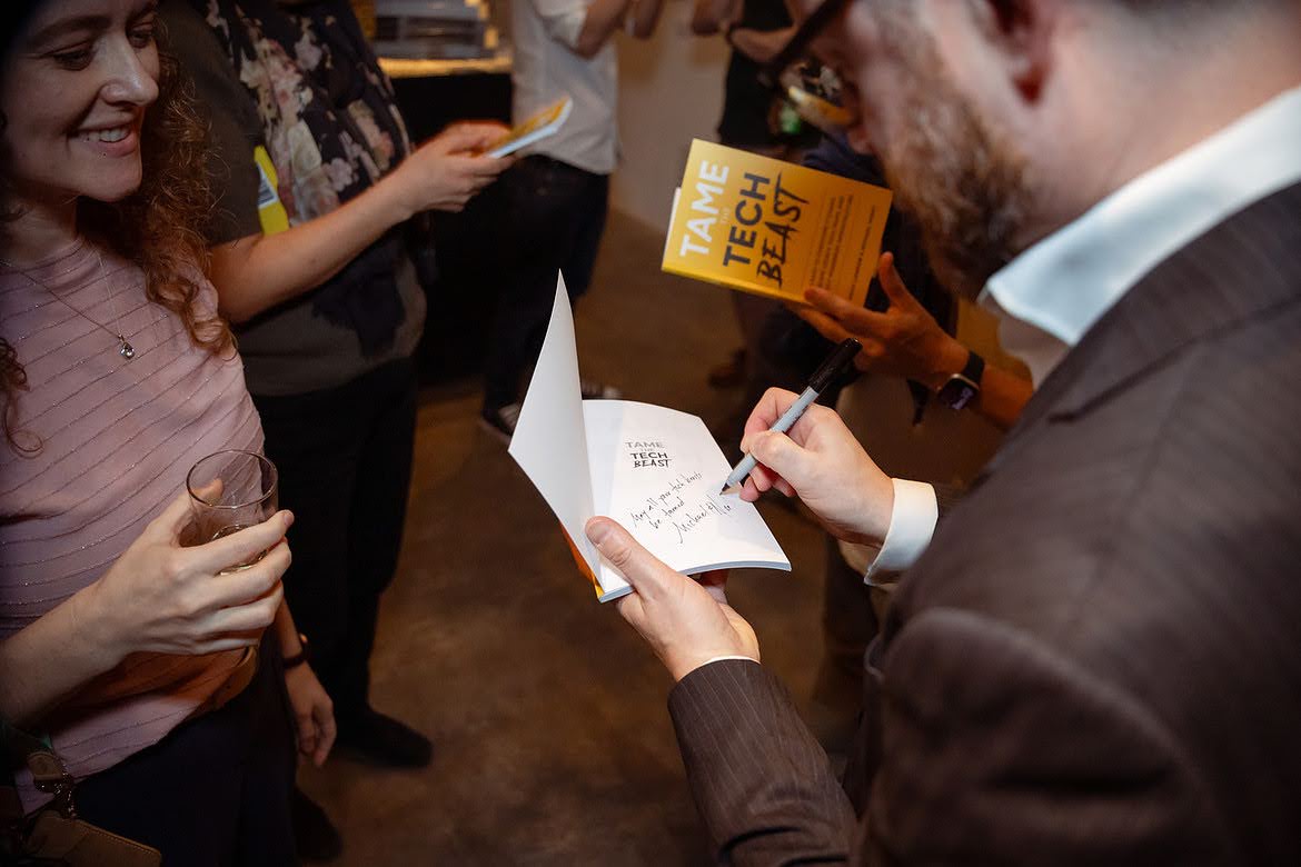 An author signing a copy of Tame the Tech Beast at the launch party while a guest looks on